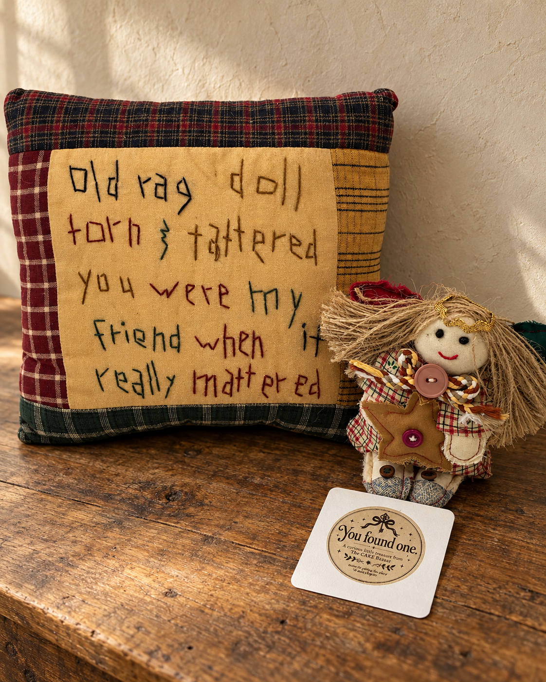 A plaid friendship pillow with an embroidered message beside a little rag angel and a "You found one" card on a wooden table.